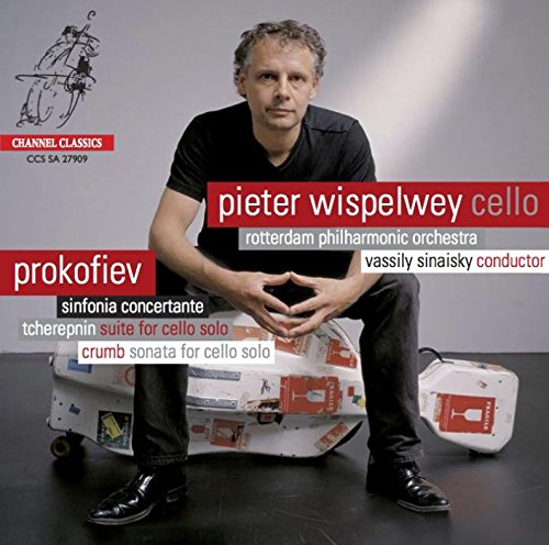 Man sitting on cello case with cello, wearing black shirt and shoes, against gray background.
