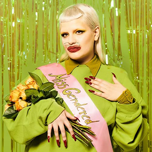 Person with drawn-on mustache, holding flowers, wearing a “Miss Gendered” sash.
