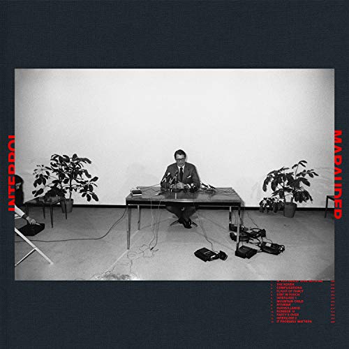 "Black and white album cover with a man at a table, surrounded by plants."