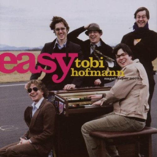 "CD cover featuring five men around a piano on an outdoor road."
