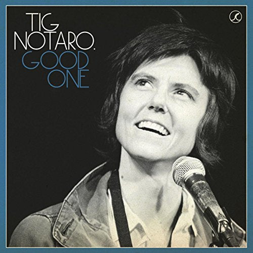 Black and white portrait of Tig Notaro smiling into a microphone on stage.
