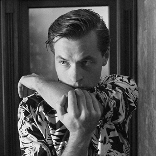 Black and white portrait of a man leaning against a door frame, wearing a patterned shirt.