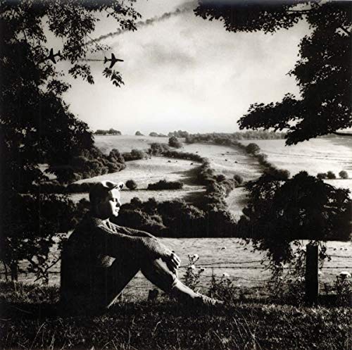 The image is a black-and-white photograph of a man sitting on the ground. #### Alt Text: Man sitting on grass, looking up at planes flying overhead in a black-and-white image.