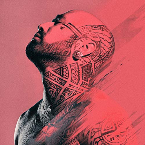 Nahko, profile view with extensive facial and neck tattoos, red background.