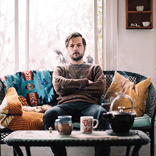 Man with beard sits in chair with tea and cushions.