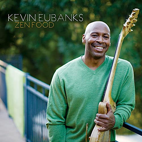 Kevin Eubanks smiles, holding a long-necked string instrument outdoors.