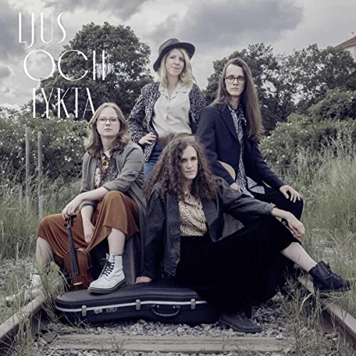 Four people with long hair pose on train tracks near trees.