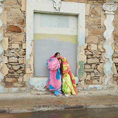 Two people in colorful, textured outfits embrace in front of a stone wall.