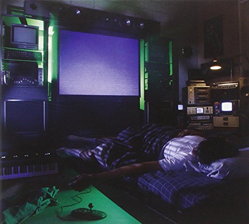 A man lies in bed surrounded by electronic equipment in a dimly lit room.