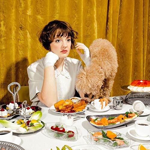 Woman with poodle and lavish food spread against a yellow curtain.