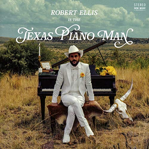 Man in white suit sits on cowhide, beside a piano, in a field.