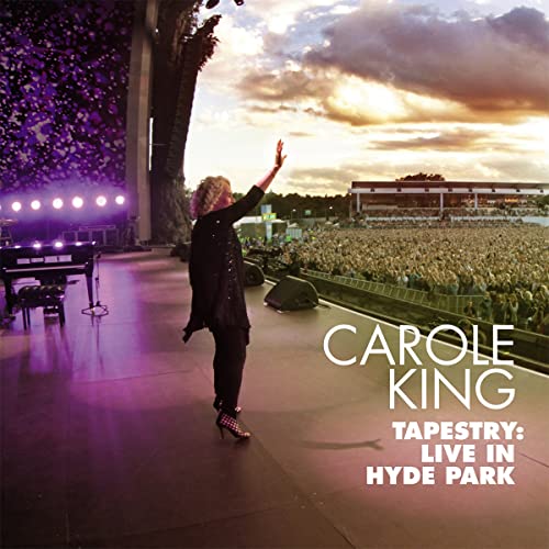 Carole King on stage at Hyde Park, with a large crowd and sunset sky.
