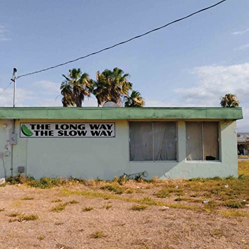 "Abandoned white building with green roof and 'The Long Way The Slow Way' sign."