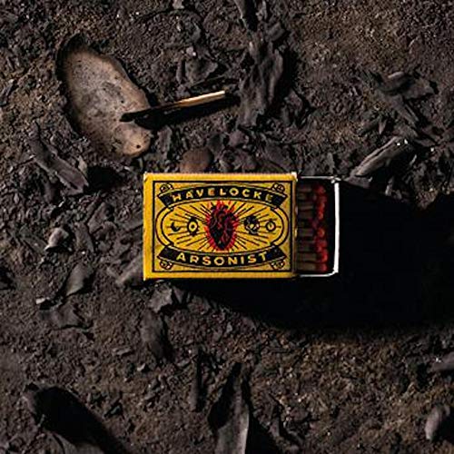 A yellow matchbox labeled "Havelocke Arsonist" on dark, charred ground with scattered debris.