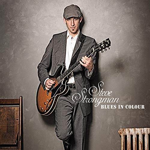 Man in gray suit and cap holding guitar, leaning against a gray wall.