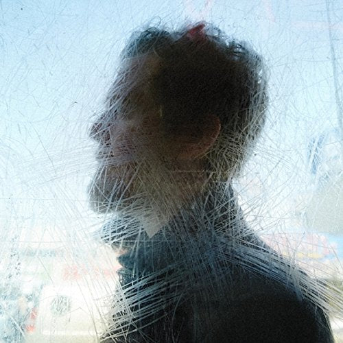 A man with a beard and dark shirt is shown through a scratched glass surface.