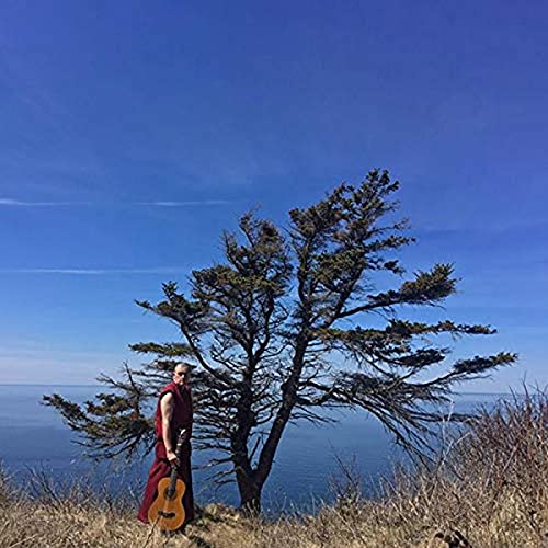 Man in red robes with a guitar stands by a tree and ocean.