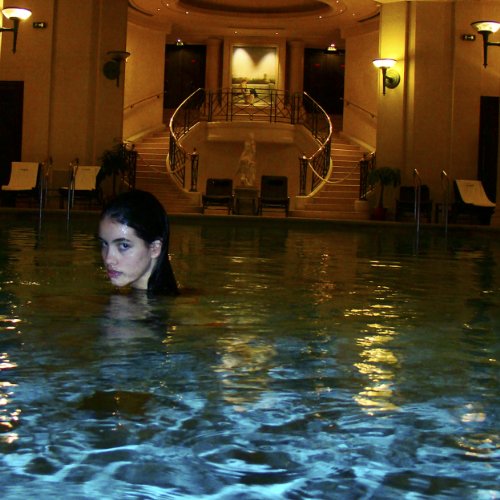 The image shows a woman in an indoor pool with a staircase and lounge chairs.