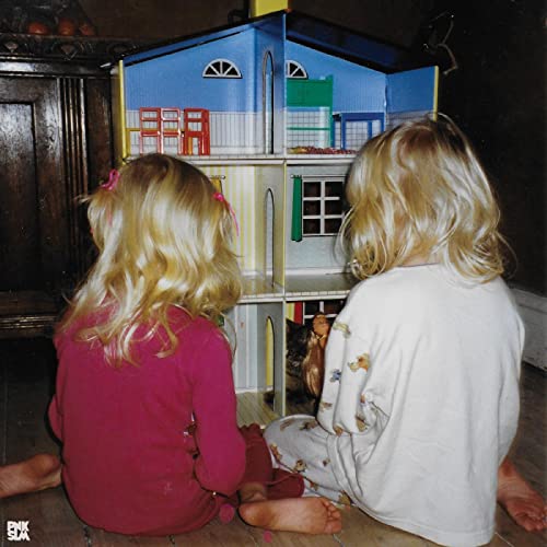 Two children with blonde hair sit facing a colorful dollhouse.