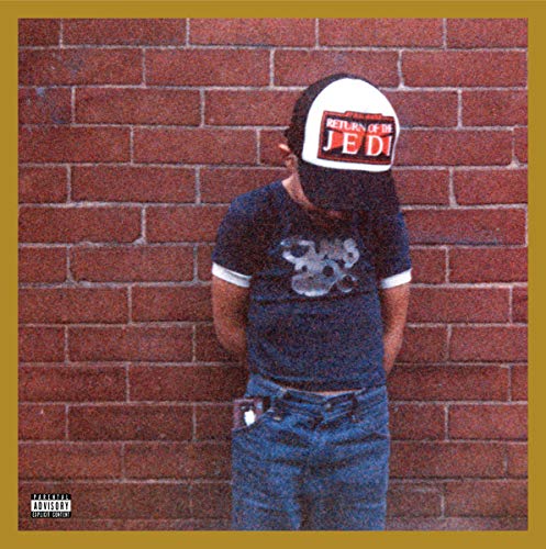 A man wearing a hat and t-shirt stands against a brick wall.
