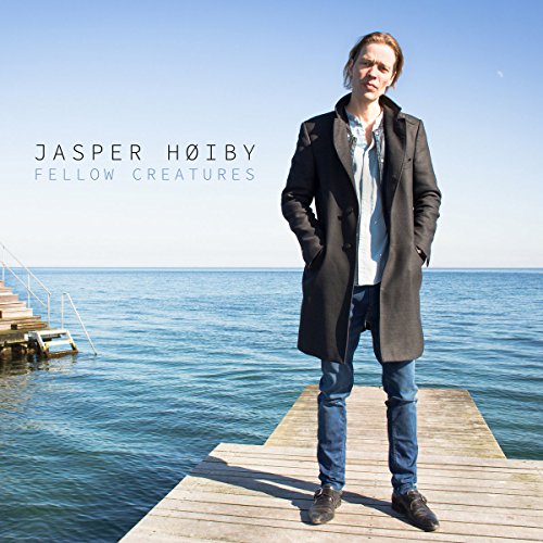 Man in dark coat and jeans stands on dock, hands in pockets, by calm blue water.