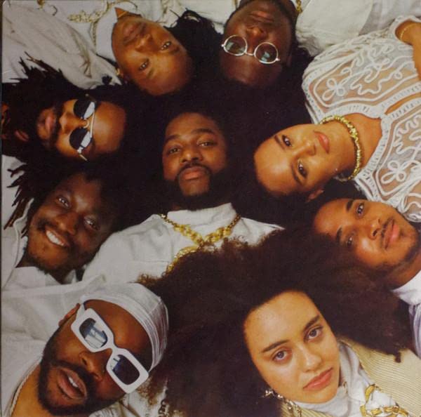 Group portrait of eight people lying in a circle, wearing sunglasses and gold chains.