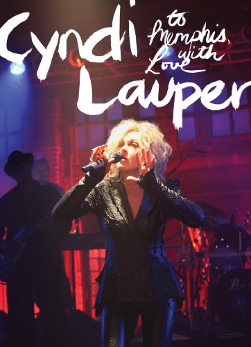 Cyndi Lauper on stage, singing into a microphone, with band members in the background.