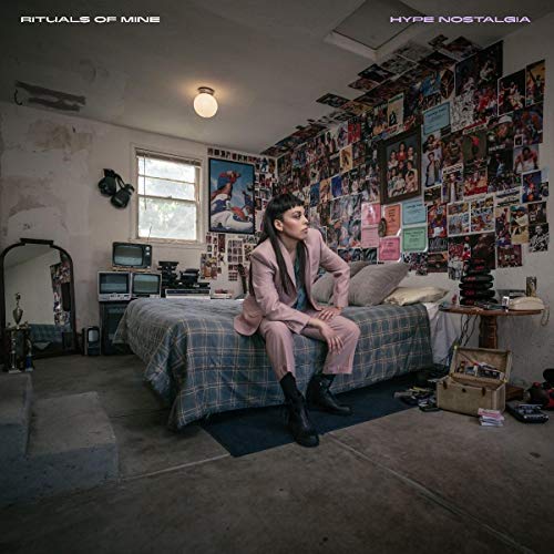 Woman in a suit sits on a bed in a cluttered room.
