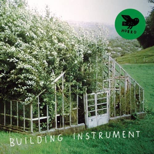 Album cover with a greenhouse, green hills, and the band name “Building Instrument”.