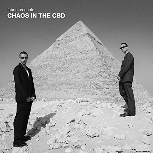 Two men in suits stand before a large, rocky pyramid.