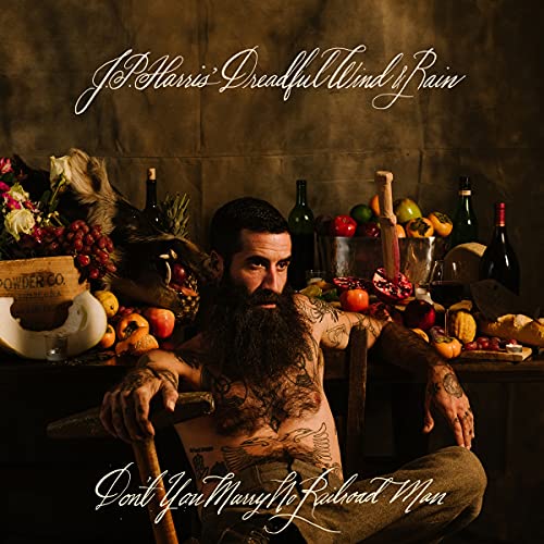 Bearded man with tattoos reclines amidst fruit, bottles, and flowers.