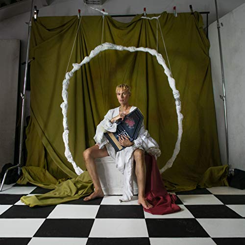 Person with album cover seated on stool, checkered floor, green backdrop.