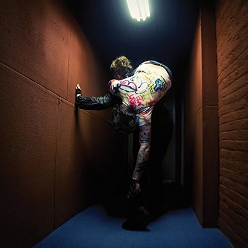 A man in a graffiti-covered jacket leans against a wall in a dimly lit hallway.