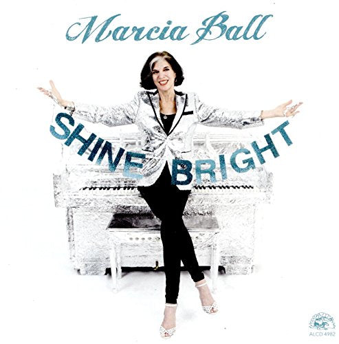 Woman standing in front of a white piano, arms outstretched, wearing a sparkly jacket.