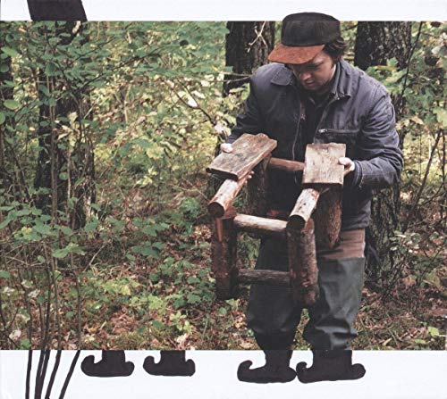 Man in cap stands with a rustic, wooden chair in a forest.