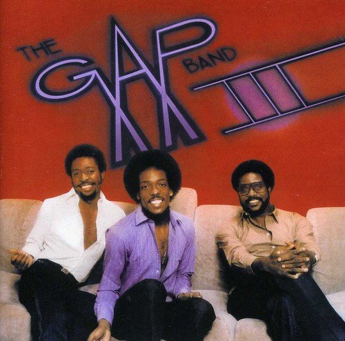 "Album cover featuring three men sitting on a couch, against a red background."