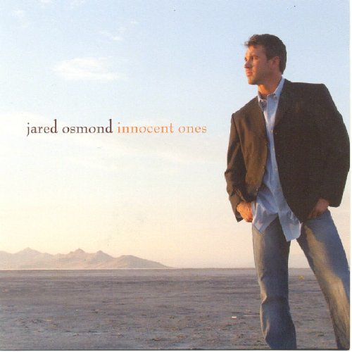 Man standing on dry land with mountains in background, wearing jacket and jeans.