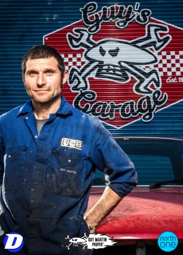 Guy Martin in blue overalls stands before a red car and garage logo.