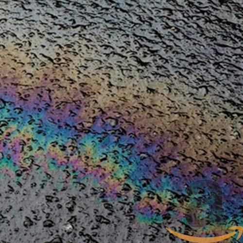The image depicts a close-up of an oil slick on water. Alt text: "Rainbow-colored oil slick on dark, textured surface with Amazon logo overlay."