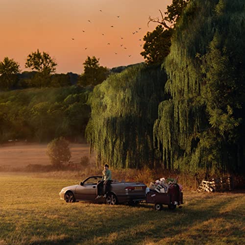 Person sitting on car with trailer in a grassy field at sunset.