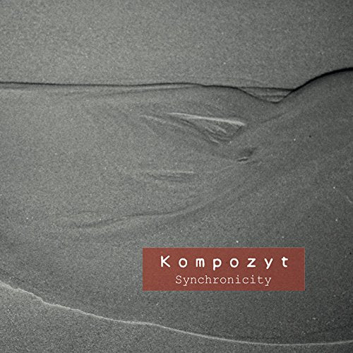The image is a CD cover with a red label. "CD cover with red label, 'Kompozyt Synchronicity', on a gray sand background."