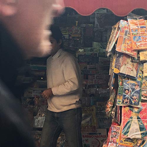 Man in white sweater stands before a cluttered record store display.