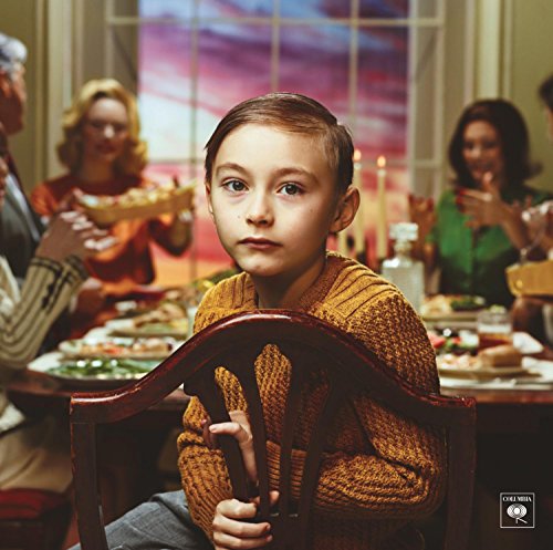 A young boy in an orange sweater sits alone at a dinner table with others.