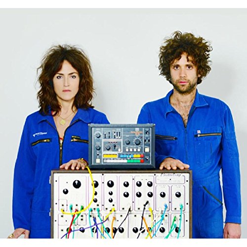 "Man and woman in blue jumpsuits stand behind electronic synthesizer equipment."