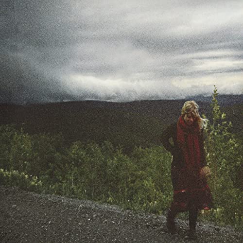Woman with red scarf stands on gravel, looking towards distant hills.