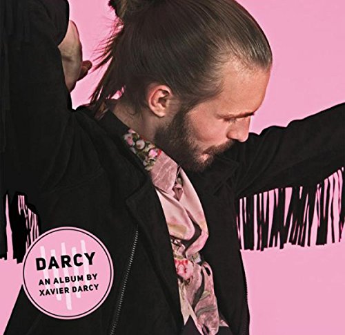 Man with dark hair and beard wearing black jacket and patterned shirt against pink background.