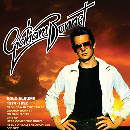 Graham Bonnet in white jacket and sunglasses against a cloudy orange sky background.