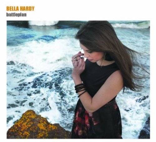 Woman in black dress stands by ocean, holding shell to her face, hair blowing.