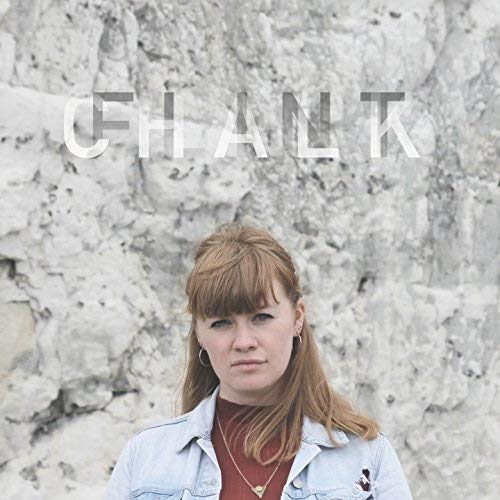 The alt text for the image is: "Woman standing in front of a white chalk wall with 'CHALK' written."