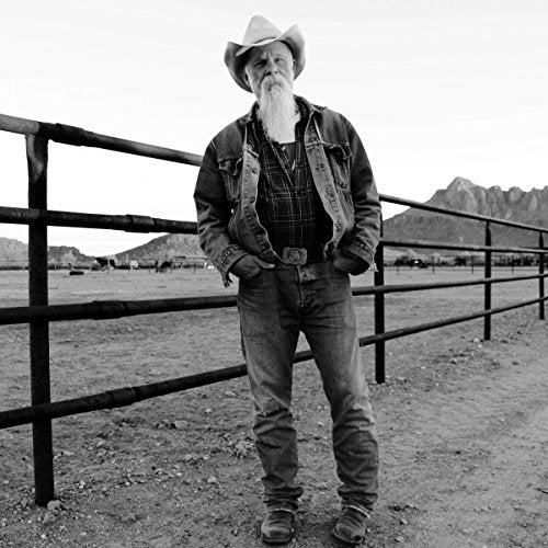 Black and white image of man wearing cowboy hat standing by a metal fence.